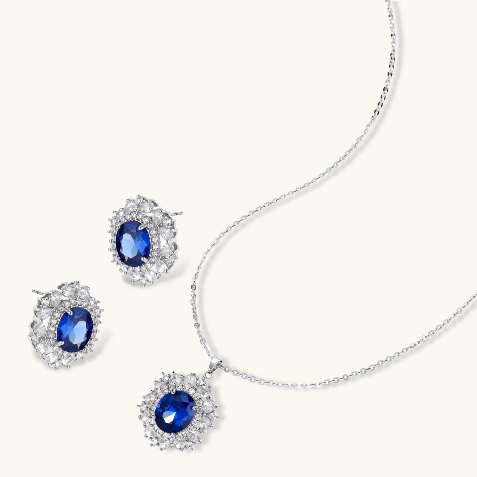White gold plated jewelry set with blue cubic zirconia centers surrounded by clear crystal halos on cream background.