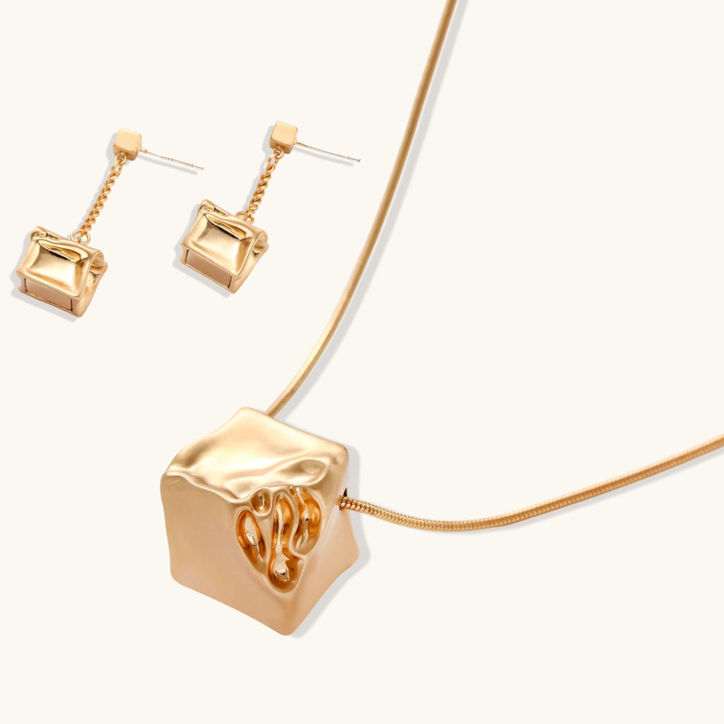 Modern geometric jewelry set featuring crumpled cube pendants in warm gold finish with chain details on beige background.