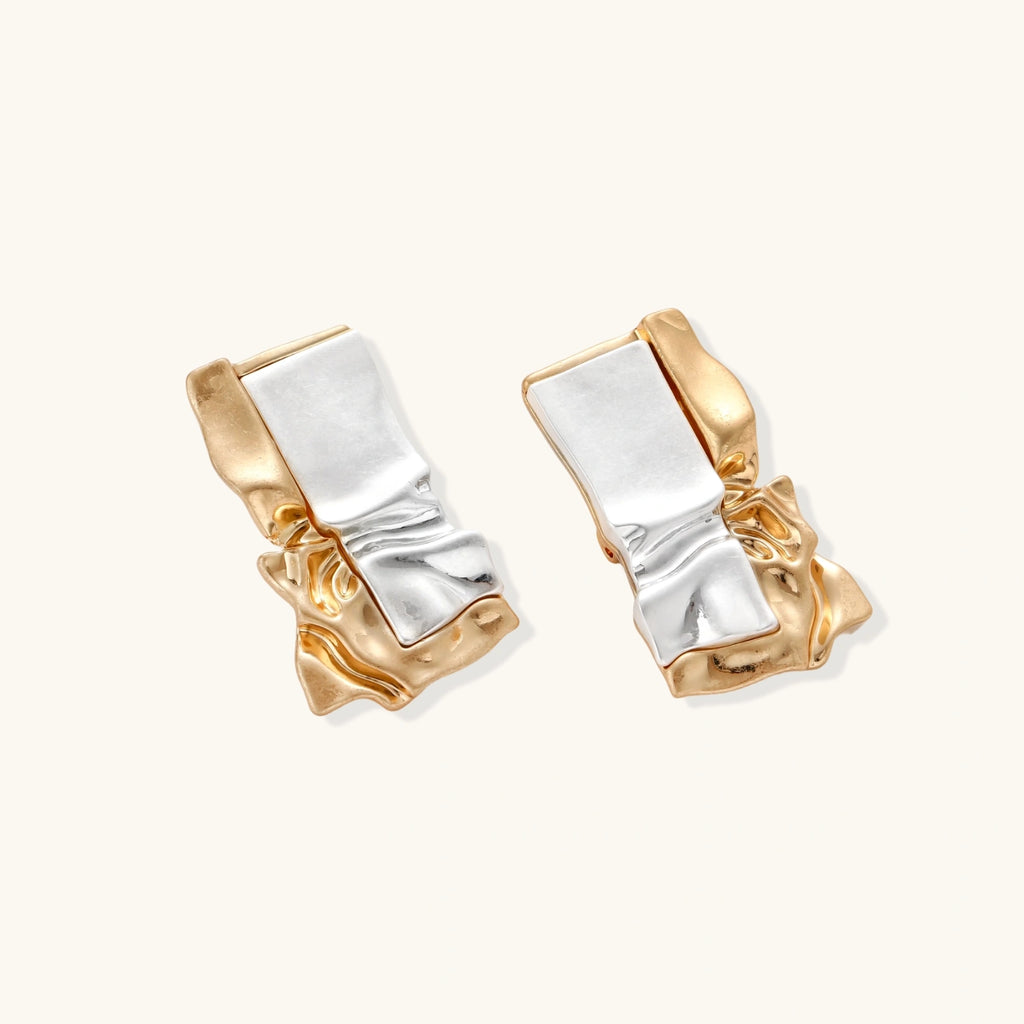 Close-up of avant-garde sculptural earrings with flowing liquid metal design in gold and silver tones on white background.