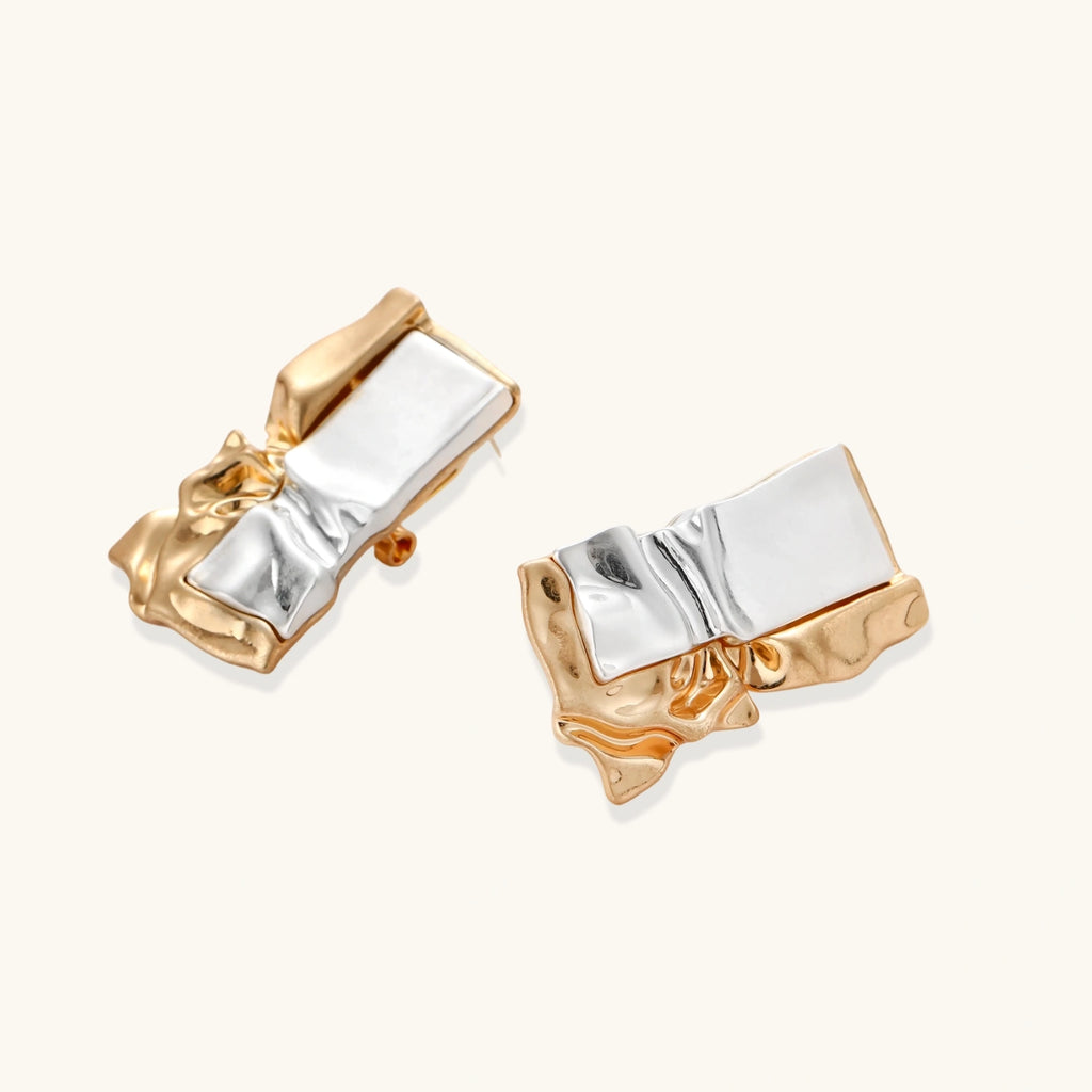 Close-up of avant-garde sculptural earrings with flowing liquid metal design in gold and silver tones on white background.