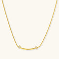 Delicate gold-plated necklace with curved bar pendant and crystal accents on snake chain against cream background.