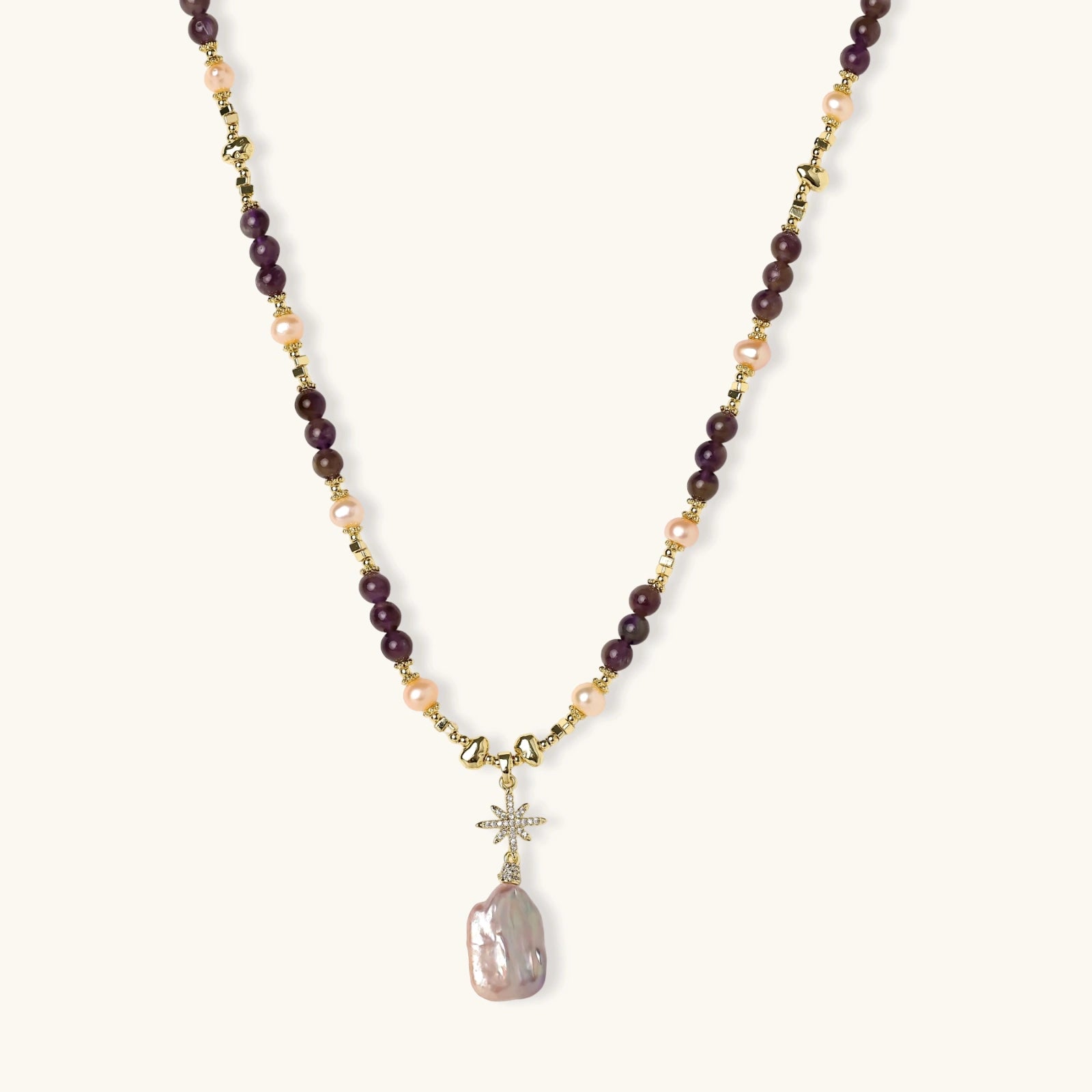 Gold necklace with iridescent baroque pearl pendant, purple amethyst beads, and crystal star charm on cream background