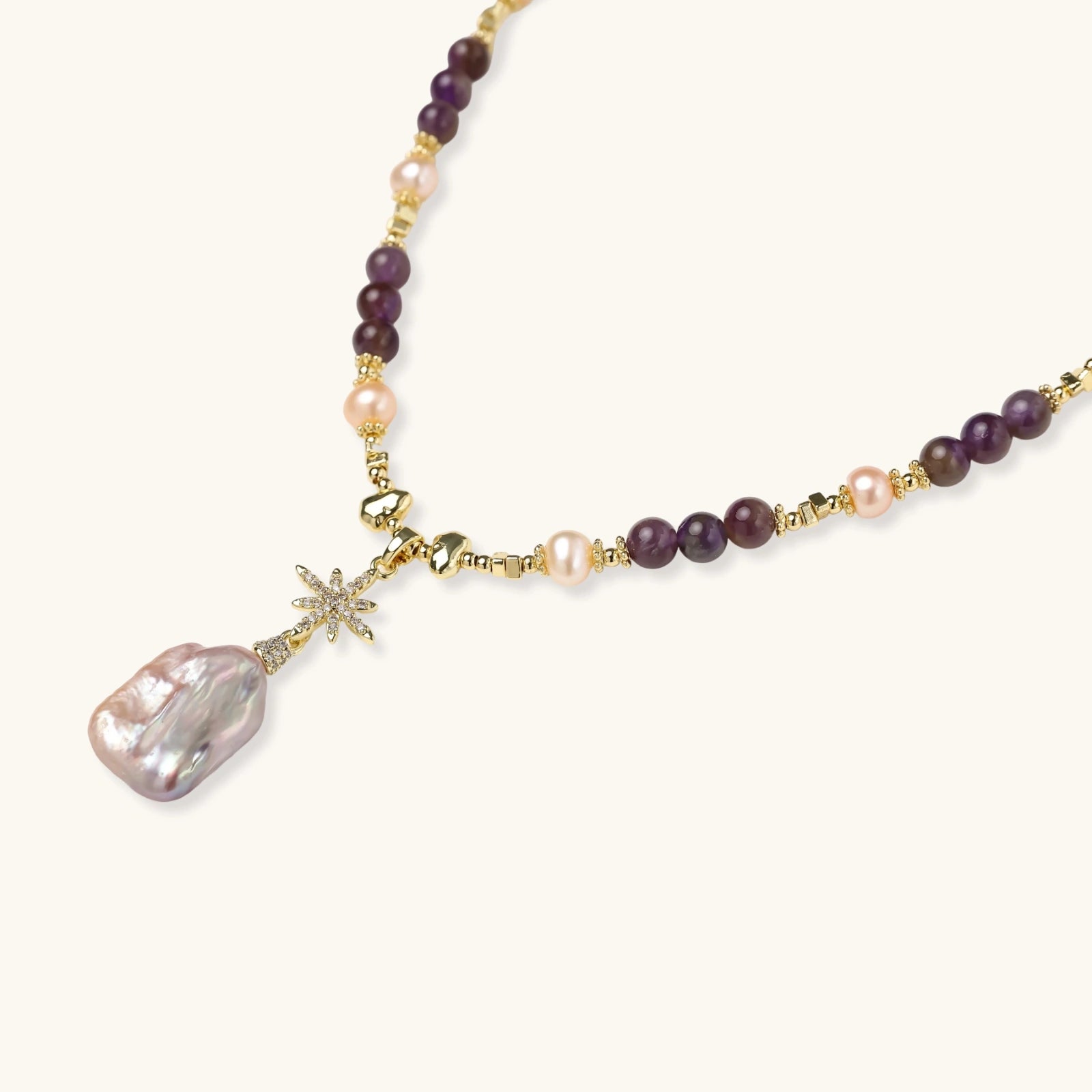 Gold necklace with iridescent baroque pearl pendant, purple amethyst beads, and crystal star charm on cream background