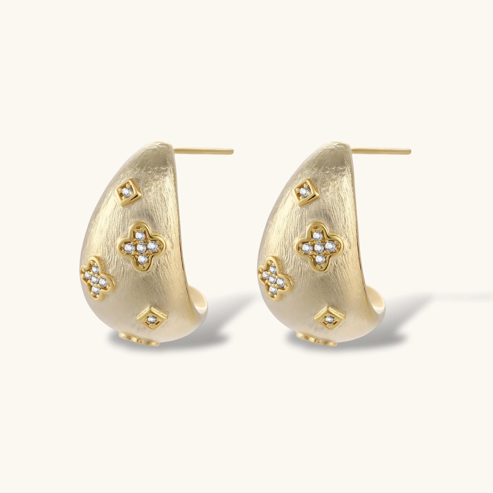 Curved gold-plated hoop earrings with four-leaf clover and diamond-shaped crystal details on white background