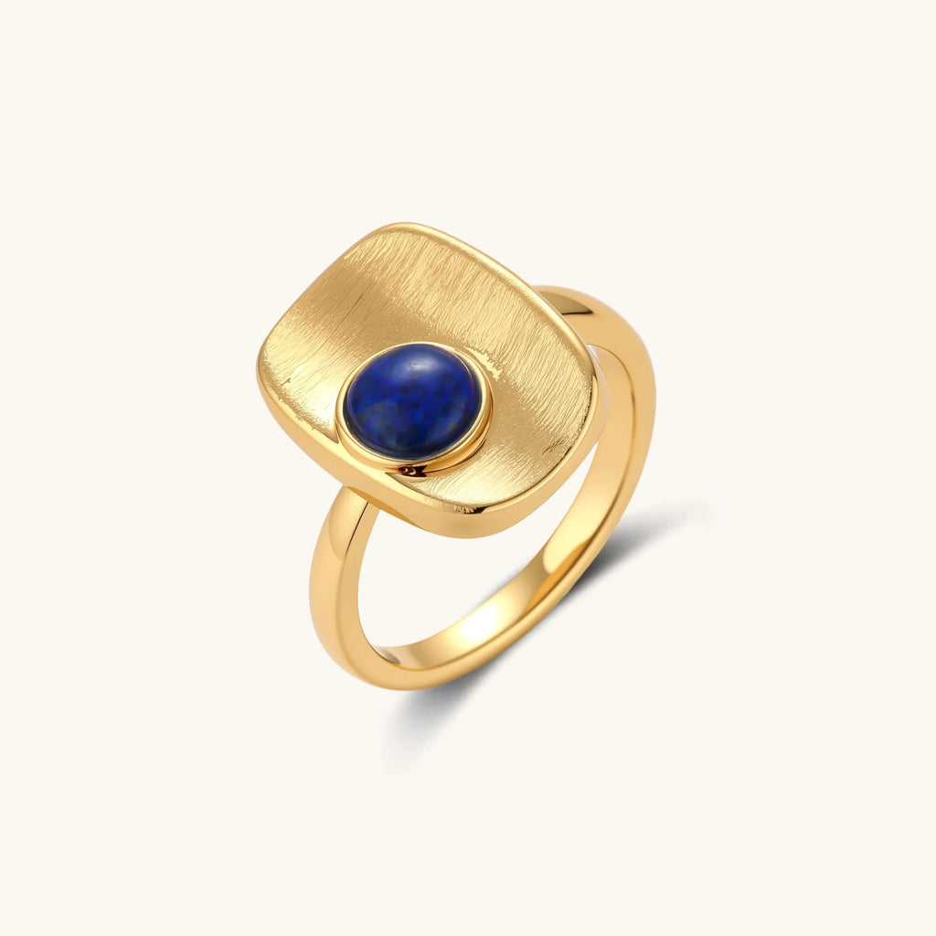 Adjustable gold-plated ring with textured square setting featuring a round blue lapis lazuli stone on cream background.